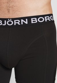Crne bokserice s elastičnim pojasom teksture na kojem piše "BJÖRN BORG" u srebrnoj boji. Tkanina je glatka s ravnim šavovima.