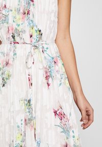 Robe blanche plissée avec imprimé floral pastel et une fine ceinture nouée à la taille, montrée des épaules aux hanches avec un bras visible.