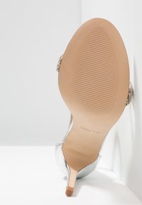 Chaussure à talon haut argentée avec un bout pointu et un talon fin. La semelle est lisse avec une texture nervurée, ornée de strass décoratifs sur la sangle.