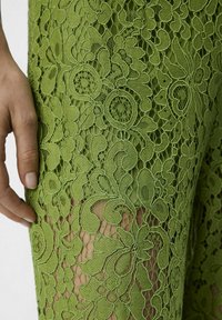 Tela de encaje verde con patrones florales y detalles intrincados. Una mano está posicionada cerca del encaje, destacando su textura y diseño.
