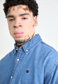 Denim overhemd met knopen in medium blauw met een kraag, heeft witte knopen en een klein logo op de linkerkant van de borst. Zachte textuur, relaxte pasvorm.