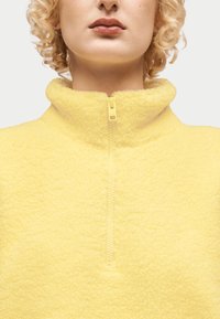 Κοντινό πλάνο ατόμου με σγουρά ξανθά μαλλιά που φοράει κίτρινο φλις μπουφάν με φερμουάρ και ψηλό γιακά.