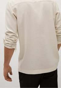 Homme portant une chemise beige à manches longues retroussées et un pantalon noir, vu de dos sur un fond uni.