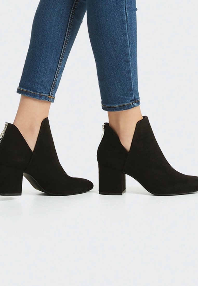 Bottines en daim noir à talon carré. Présentent un design découpé sur les côtés, une texture contrastante et un accent métallique à l'arrière.