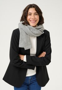 Écharpe en laine grise enroulée autour du cou, portée avec un blazer noir et une chemise blanche. Texture douce, forme rectangulaire, motif subtil. Look décontracté.