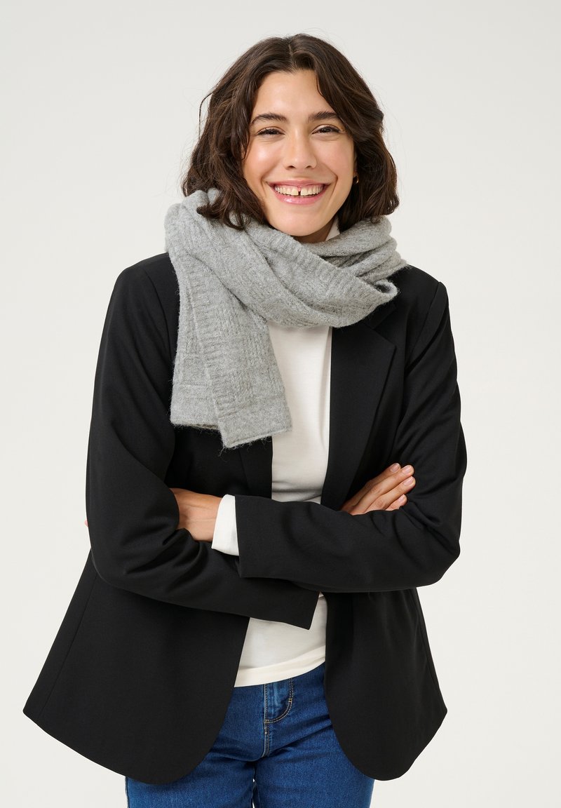 Écharpe en laine grise enroulée autour du cou, portée avec un blazer noir et une chemise blanche. Texture douce, forme rectangulaire, motif subtil. Look décontracté.