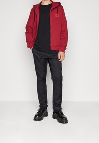 Červená bunda s kapucí na zip, černé tričko a černé kalhoty. Boty s tlustou podrážkou. Jednoduchý design, bavlněná textura, minimální akcenty. Jednoduché pozadí.