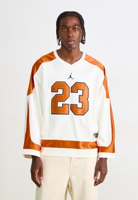 Young man with braided hair wearing white and orange sports jersey with number 23 and beige pants, standing against plain background.