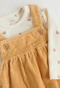 Senf-farbene Cord-Dungaree mit Rüschensaum und Knopfakzenten, über einem cremefarbenen Langarmshirt mit kleinen Drucke von braunen Bären.