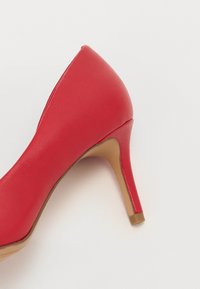 Chaussure à talon haut rouge avec un bout pointu, en cuir synthétique lisse, un talon fin et une semelle texturée marron.