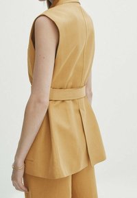 Ärmlös senapsgul väst med insydd midja, fö featuring en utsvängd bakdel, slagkrage och sidofickor. Slät tyg med en ren finish.