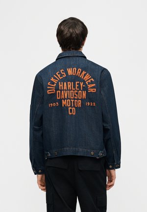 Person som har på seg en mørk denimjakke med oransje tekst "Dickies Workwear Harley-Davidson Motor Co" på ryggen, kombinert med svarte bukser.