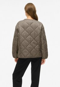 Femme de dos portant une veste matelassée taupe avec un motif de couture en ondulation et un pantalon noir sur fond blanc uni.