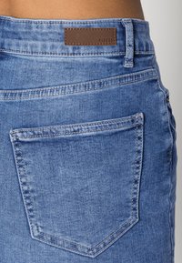 Jean en denim bleu délavé avec une étiquette en cuir marron cousue à la taille et une poche arrière unique de forme classique.