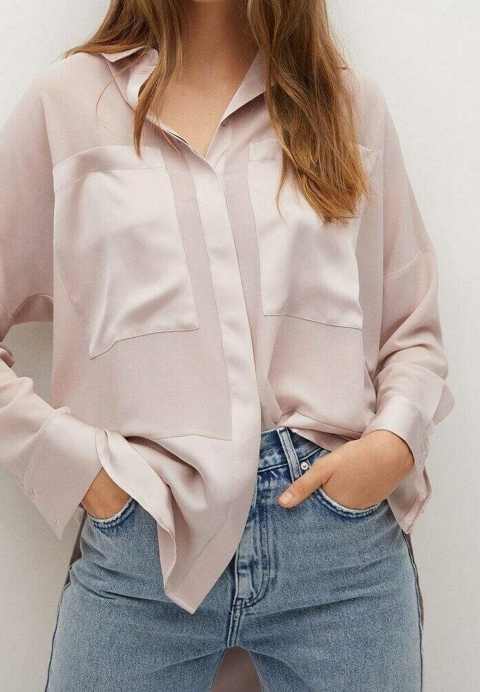 Femme portant une chemise ample en satin rose clair avec des poches à l'avant, rentrée dans un jean bleu délavé taille haute, mains dans les poches.