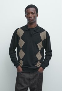 Zwarte trui met lange mouwen, beige ruitpatronen, een kraag en een textuurrijke stof. Model draagt donkergrijze broek.