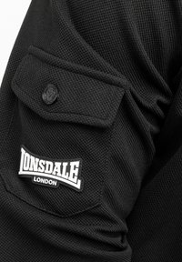 Fekete textúrájú anyag, amelyen egy kiemelkedő zseb található fekete gombbal. Tartalmaz egy fekete-fehér logót a "Lonsdale London" felirattal.