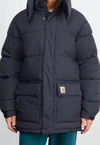 Persona vistiendo una chaqueta de invierno acolchada negra con bolsillos con solapa y una capucha, de pie contra un fondo liso.