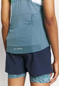Top atletico color teal con accenti bianchi e verdi, presenta arricciature e un orlo testurizzato. Abbinato a pantaloni corti color navy e strati sottostanti stampati.