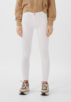 Personne portant un jean slim blanc, des baskets beiges et marron épaisses, et un pull en maille beige avec des boutons, debout sur un fond blanc.