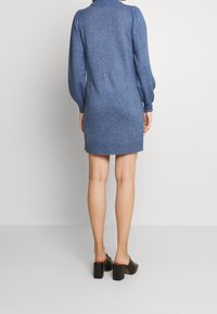 Abito in maglia blu con collo alto, maniche a sbuffo e silhouette aderente, che termina sopra il ginocchio. Abbinato a scarpe con tacco cubano nere.