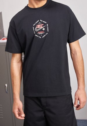 Mann trägt schwarzes Nike T-Shirt mit rotem und weißem rundem Logo mit dem Schriftzug „Sole Energy, Sole Synergy“ in einer Innenumgebung.