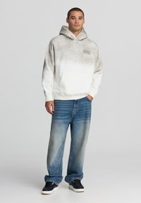 Felpa oversize bianca con gradiente grigio, dotata di tasca frontale e logo. Indossata con jeans azzurri larghi e sneakers nere.