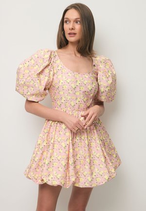 Vrouw met stijl bruin haar, die een korte roze jurk draagt met een geel bloemenpatroon en opgeblazen mouwen, staand tegen een effen achtergrond.