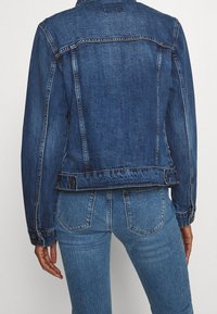 Jeansowa kurtka w ciemnym niebieskim kolorze o klasycznym kroju, z przeszyciami i zapięciem na guziki u dołu. Widok z tyłu pokazuje długie rękawy i dopasowaną sylwetkę.