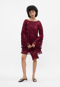 Καφέ φόρεμα με λουλουδάτη κεντημένη διακόσμηση, μακριά μανίκια τύπου καμπάνας. Το μοντέλο κρατάει μία τσάντα με αστεράκια και φοράει μαύρα σανδάλια με μικρό τακούνι.