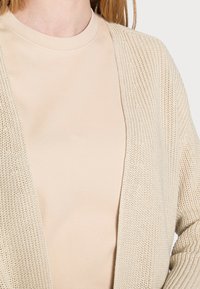 Beige offene Strickjacke über einem hellbeigen Rundhalsausschnitt-T-Shirt, das ein strukturiertes Muster und eine lässige Passform zeigt.