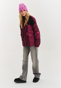Magentová pufferek s prešívanými čiernymi akcentmi, so zipsami a kvetinovými vzormi. V kombinácii so šedými širokými džínsami a čiernymi bezšnúrovými topánkami.
