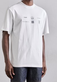 Camiseta de algodón blanca con cuello redondo, que presenta logotipos gráficos grises en la parte delantera. Corte holgado y mangas cortas.