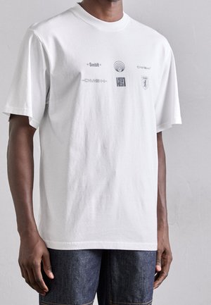 Camiseta de algodón blanca con cuello redondo, que presenta logotipos gráficos grises en la parte delantera. Corte holgado y mangas cortas.