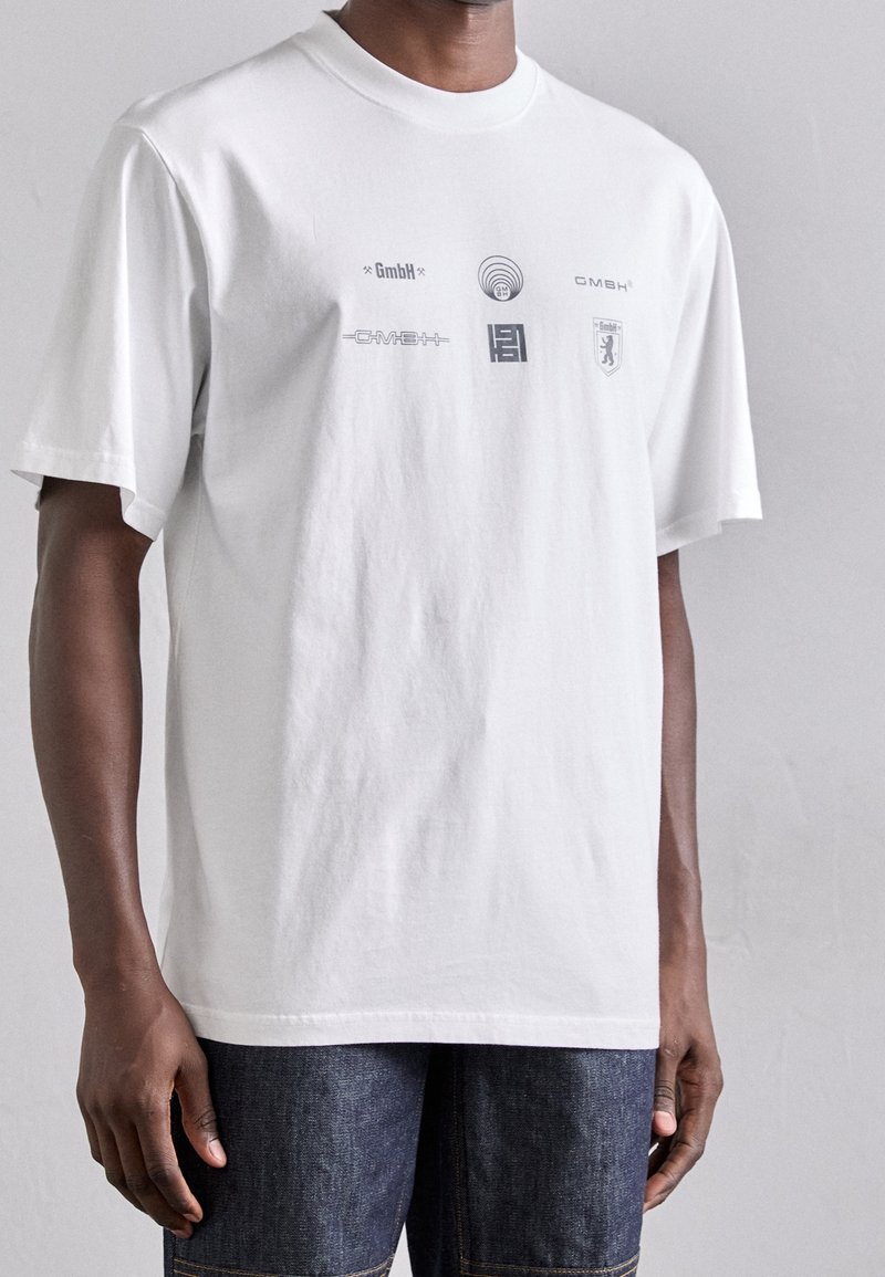 Camiseta de algodón blanca con cuello redondo, que presenta logotipos gráficos grises en la parte delantera. Corte holgado y mangas cortas.