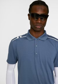 Pikepaita sinisestä kankaasta, jossa valkoiset korostusraidat hartioilla. Klassinen kaulus ja kolme nappia napituksessa. Aurinkolasit täydentävät ilmeen.