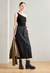 Black one-shoulder bodysuit, high-waisted black denim skirt, and black platform ankle boots. Holding a tan coat. Smooth texture, minimal design.