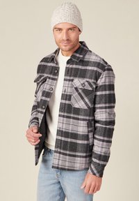 Zwart en grijs geruite flanellen shirt met twee borstzakken, knoopsluiting aan de voorkant en een grijze gebreide beanie. Witte binnenlaag en lichtblauwe spijkerbroek.