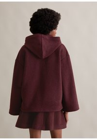 Bordowa kurtka z kapturem o fakturowanym wykończeniu, szerokich rękawach i krótkim kroju. Modelka ma na sobie pasującą rozkloszowaną spódnicę.
