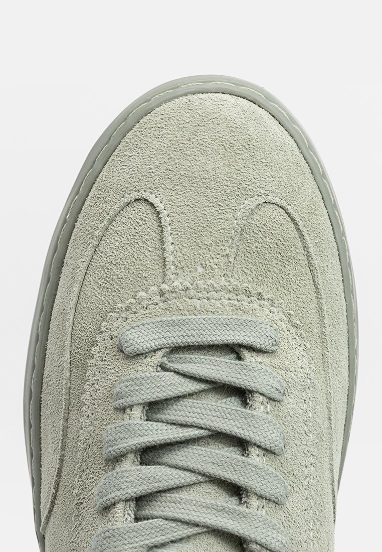 Baskets en daim vert clair avec une tige texturée, bout rond et lacets plats. Détail de couture subtil le long du bord de la chaussure et des lacets.