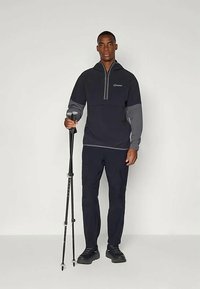 Sudadera de forro polar en azul marino oscuro con acentos grises, diseño de medio cierre, combinada con pantalones oscuros y un bastón de trekking, llevada con zapatos negros.