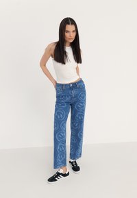 Jeans de mezclilla azul claro con un corte relajado, que presentan un patrón repetido de caras sonrientes. Combinados con una camiseta sin mangas blanca y zapatillas negras.