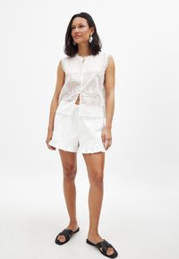Hvid ærmeløs bluse med blomsterbroderi og knappelukning, parret med hvide rynkede shorts og sorte slidssandaler.
