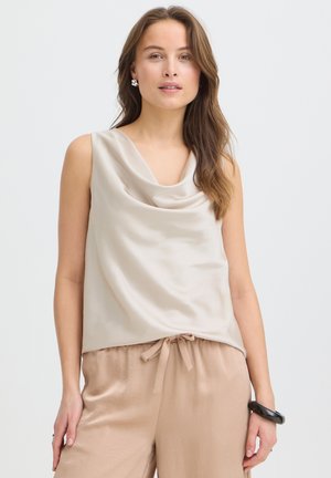 Femme aux longs cheveux bruns portant un haut drapé sans manches de couleur crème et un pantalon en satin beige, accessoirisée de boucles d’oreilles argentées et d’un bracelet noir.