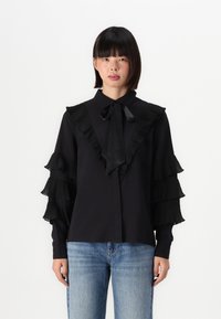 Maglietta nera con dettagli a balze plissettate su maniche e colletto. Presenta un fiocco legato al collo e una chiusura a bottoni davanti. Indossata con jeans blu.
