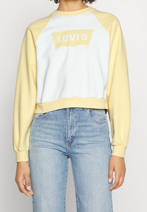 Person trägt gelb-weißes Levi's Sweatshirt und hellblaue, hoch taillierte Jeans, steht mit entspannten Armen an den Seiten.