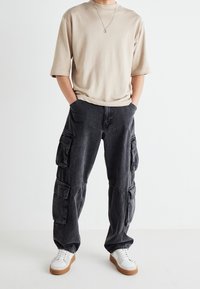 Beige oversized t-shirt med korta ärmar i kombination med mörkgrå cargobyxor med flera fickor, avslutat med vita sneakers och ett enkelt halsband.