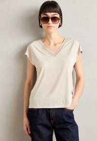Beige V-hals T-shirt met korte mouwen, voorzien van een gestructureerde rand langs de halslijn, gecombineerd met donkerblauwe spijkerbroek.