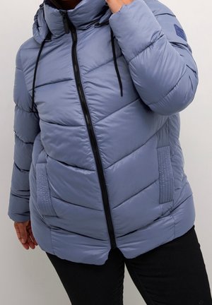 Person trägt eine hellblaue gesteppt gefütterte Jacke mit schwarzem Reißverschluss und Kordeln, kombiniert mit schwarzer Hose vor einfachem Hintergrund.