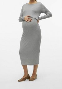 Grå långärmad gravidklänning med en tight silhuett, något rundad nederkant och mjuk ribbad textur. Buren med bruna platta skor.